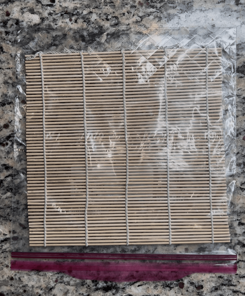 A bamboo sushi mat wrapped in plastic, lying on a granite countertop, with a pink zip-lock bag positioned underneath.