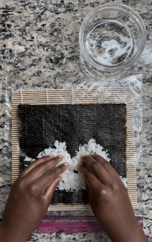 Hands spreading sushi rice on a sheet of nori placed on a bamboo mat, with a bowl of water nearby.