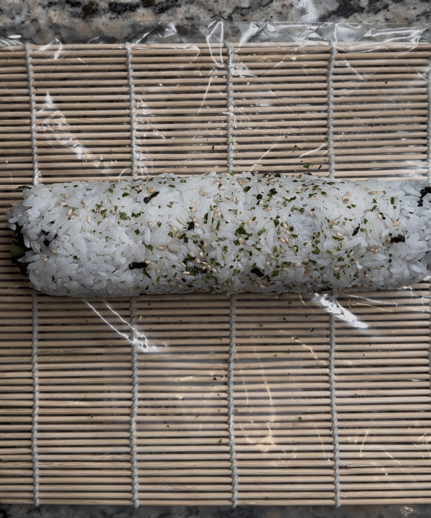 A sushi roll on a bamboo mat, wrapped in plastic, topped with sesame seeds and dried seaweed.
