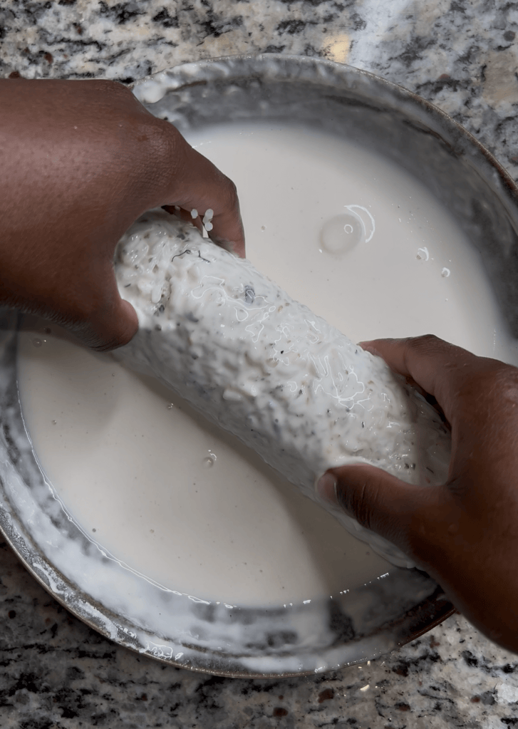 Hands holding a log-shaped food item submerged in a bowl of creamy liquid, set on a granite countertop.