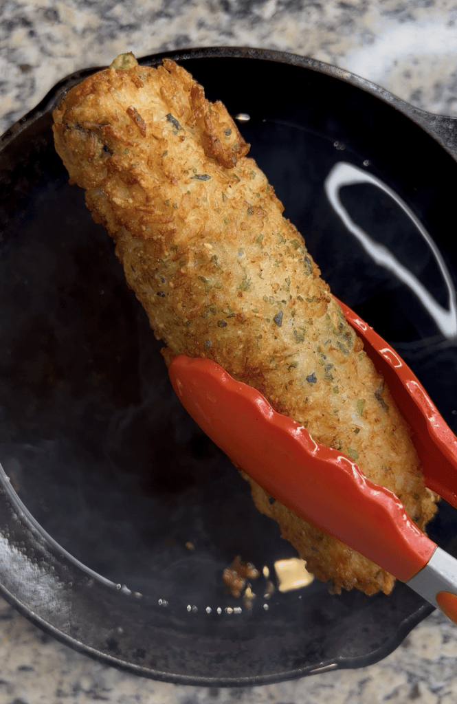 A piece of fried food being lifted with red tongs from a cast iron skillet, surrounded by steam.