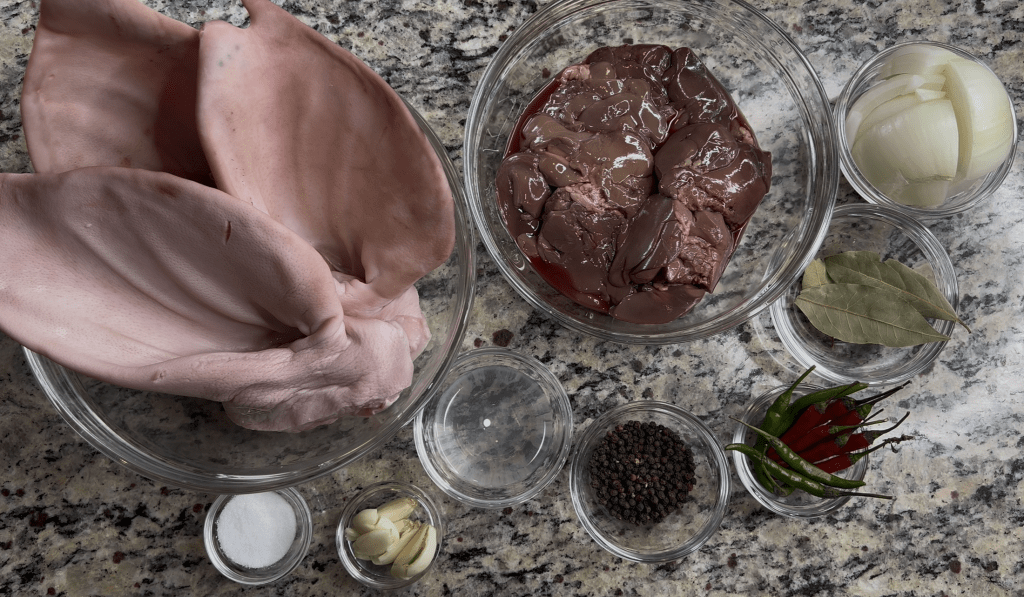 A flat lay of various ingredients for cooking, including pig's ears, liver, onions, garlic, salt, bay leaves, black pepper, and green chilies arranged in clear bowls on a granite countertop.