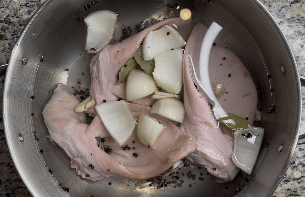A pot containing pig trotters submerged in water, with sliced onions, bay leaves, and peppercorns visible.