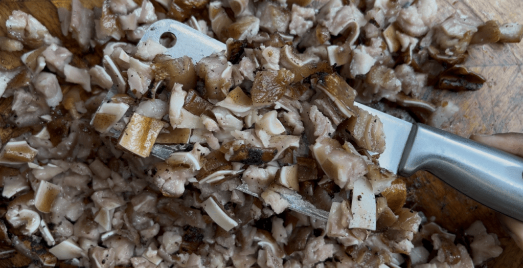 Chopped pieces of pork fat and skin on a wooden cutting board, with a knife resting on top.