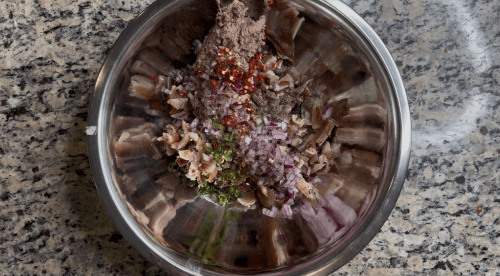 A stainless steel mixing bowl filled with various chopped ingredients, including onions, herbs, and spices, all combined for a recipe preparation.