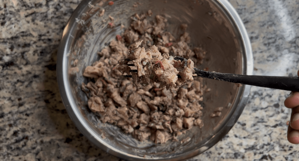 A bowl containing a mixture of shredded meat, possibly chicken or tuna, being stirred with a fork against a granite countertop.