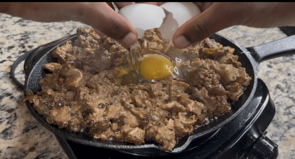 A person cracking eggs into a skillet filled with meat and vegetables on a countertop.