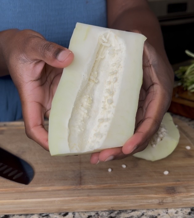A person holding a sliced cucumber that reveals the inner seeds and flesh, with a wooden cutting board and knife in the background.