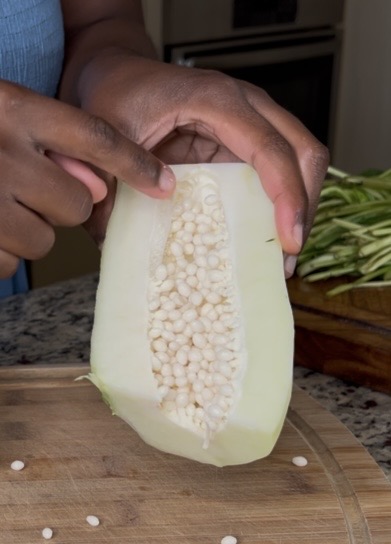A close-up of a hand holding a halved melon, showing the seeds inside, with a wooden cutting board and green vegetable strips in the background.