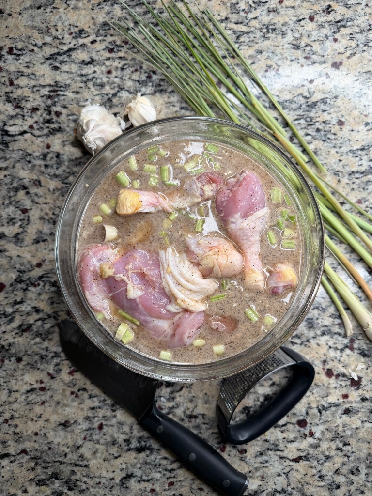 A glass bowl containing marinated chicken pieces with chopped garlic and scallions, surrounded by fresh garlic cloves and stalks of green onions on a granite countertop.