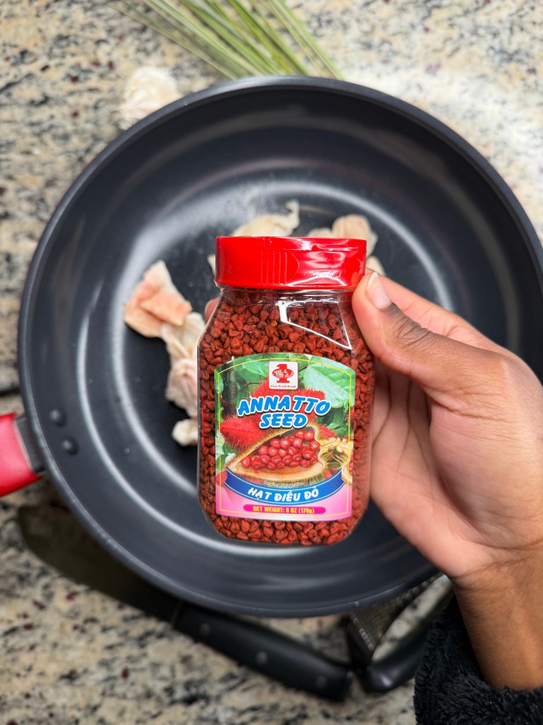 A hand holding a jar of annatto seeds over a non-stick frying pan with pieces of chicken and green herbs in the background.