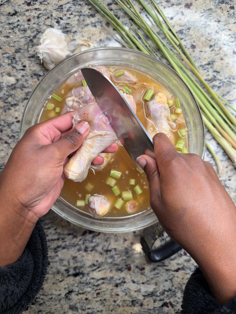 A pair of hands holding a chicken wing over a bowl of broth with chopped green onions and garlic, preparing for cooking.