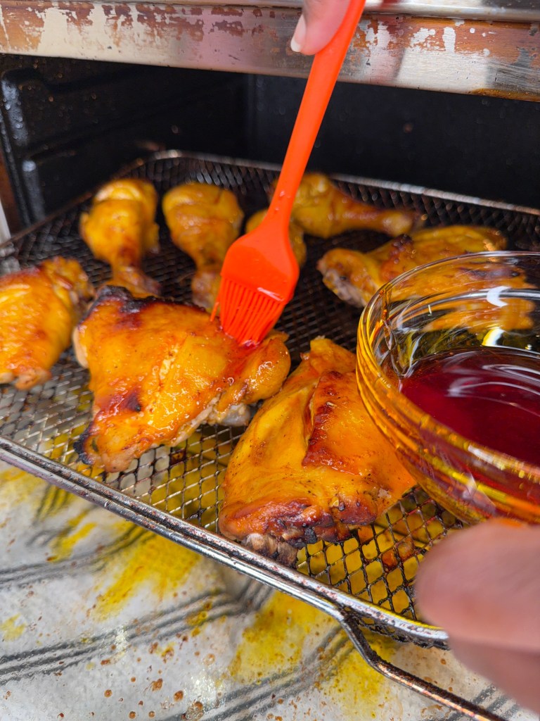A close-up of grilled chicken wings in a wire basket, being brushed with a bright orange marinade using a silicone brush, with a small bowl of sauce nearby.