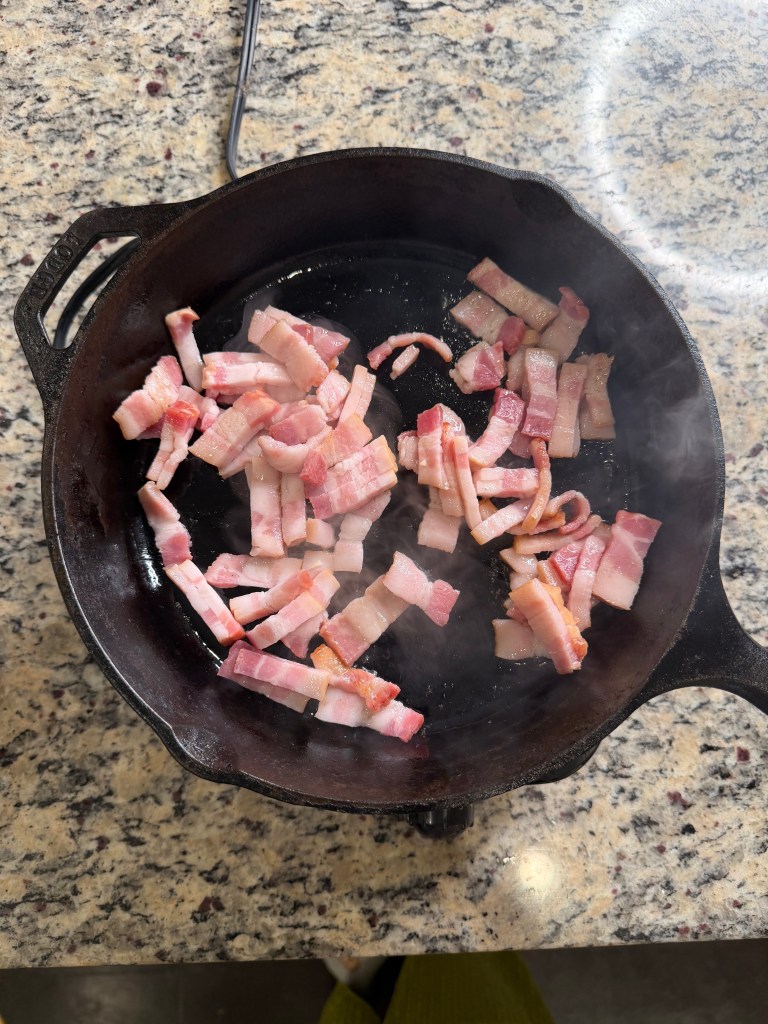 Bacon pieces cooking in a cast iron skillet on a granite countertop.