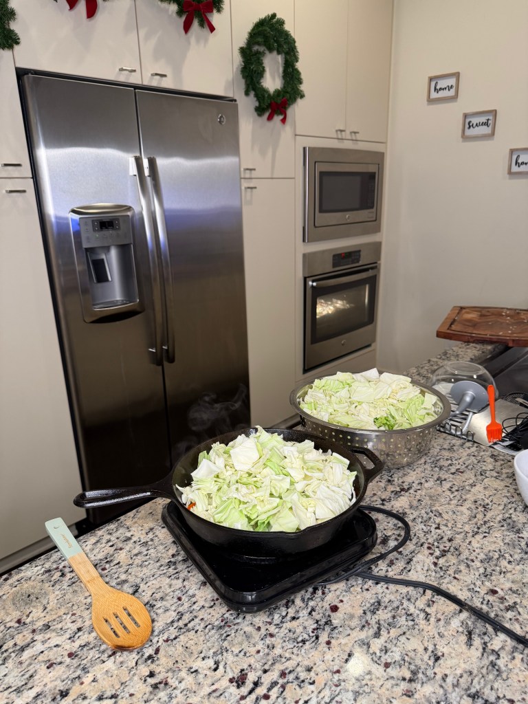 A kitchen scene featuring a pot of chopped cabbage on a stovetop, a bowl of cabbage in a colander, a wooden spoon, and modern kitchen appliances, with holiday wreaths decorating the cabinets.