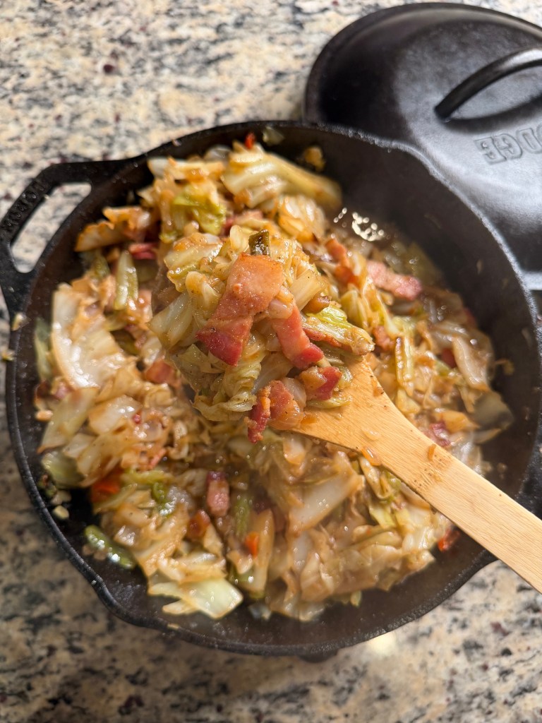 A wooden spoon lifting a mixture of sautéed cabbage and bacon in a cast iron skillet on a granite countertop.