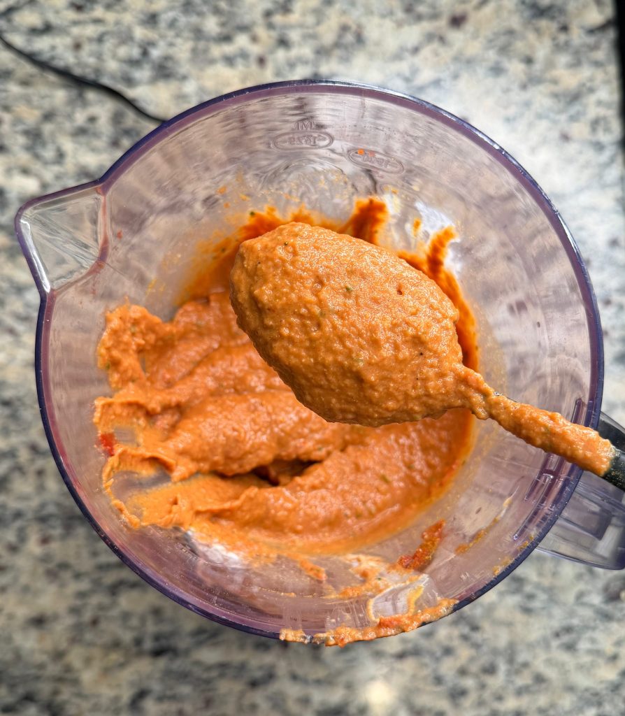 A close-up view of blended orange sauce or dip in a transparent blender jug with a spoonful of the mixture.