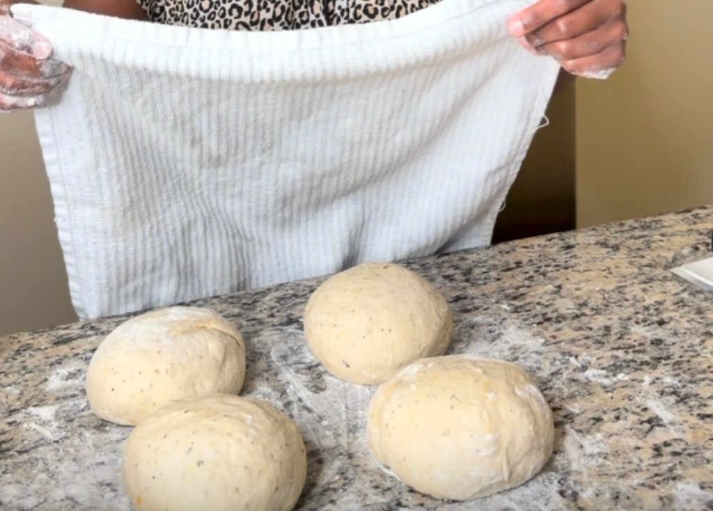 Person holding a towel in front of four portions of pizza dough on a countertop.