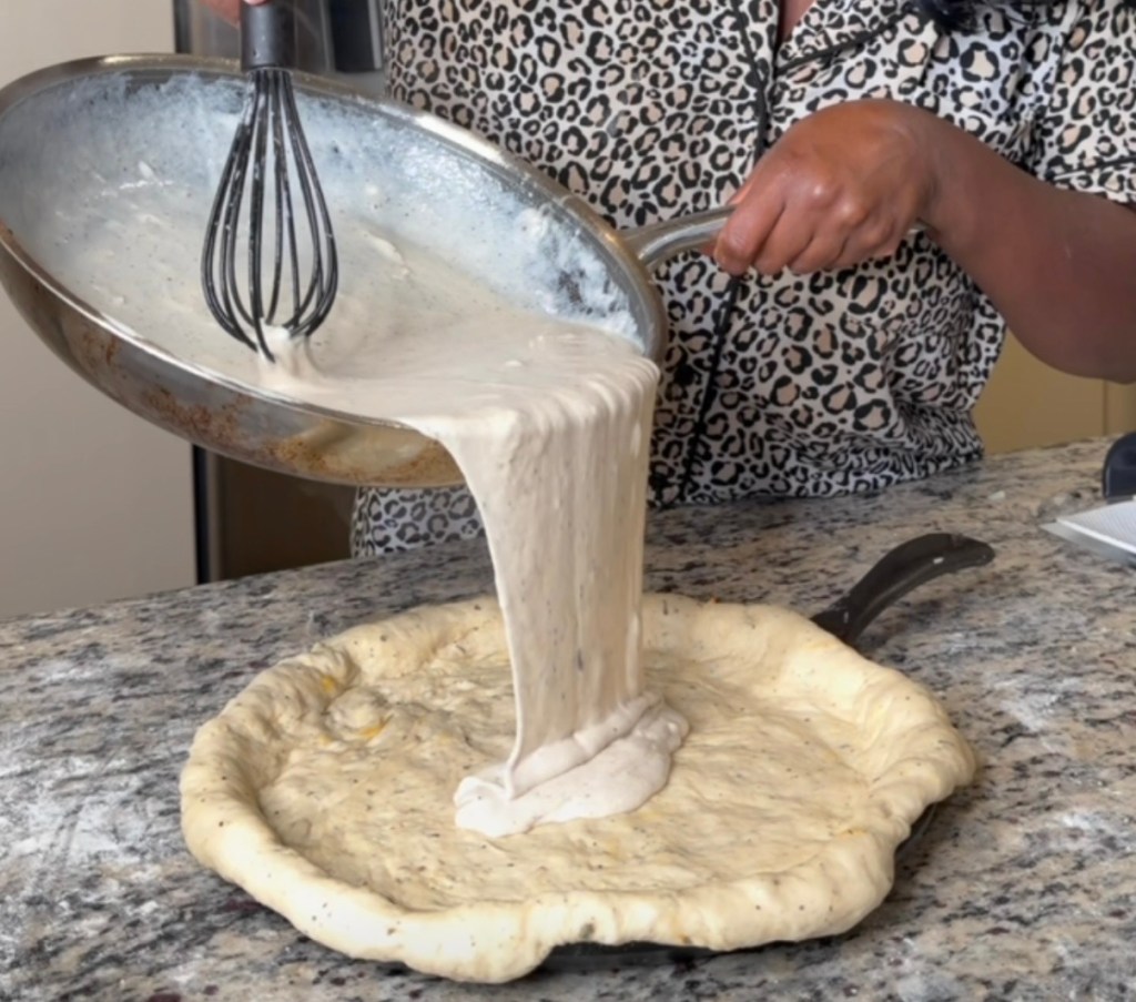Neisbae pouring creamy white sauce over a raw pizza dough crust on a countertop.