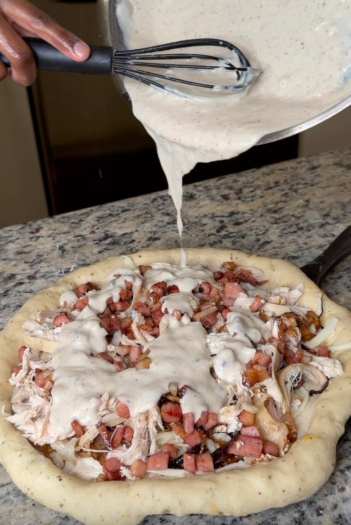 Neisbae pouring creamy white sauce over a pizza topped with shredded chicken, ham, and onions, set on a granite countertop.