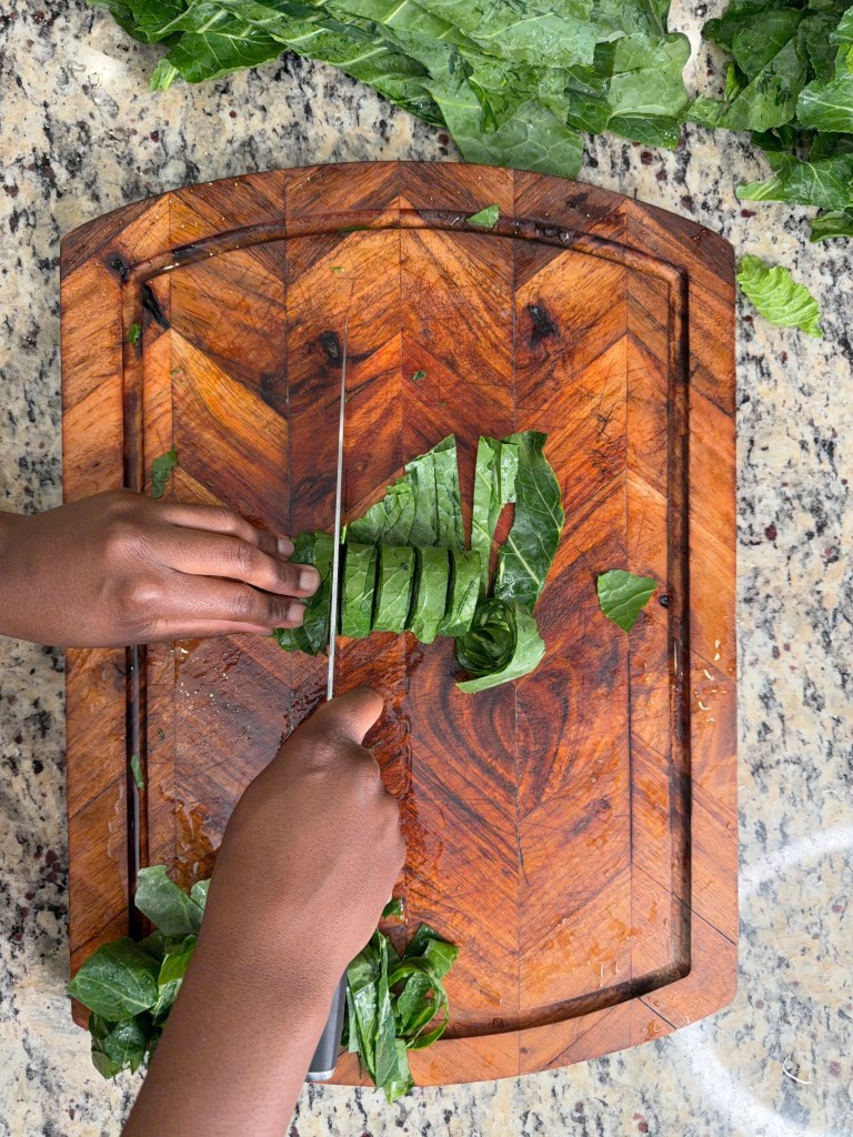 A person chopping leafy greens on a wooden cutting board with a knife, surrounded by more leafy greens.