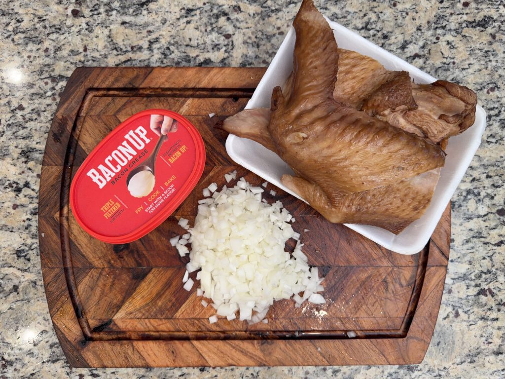 A wooden cutting board with chopped onions, a container of bacon grease labeled 'Bacon Up', and cooked chicken wings on a white plate.