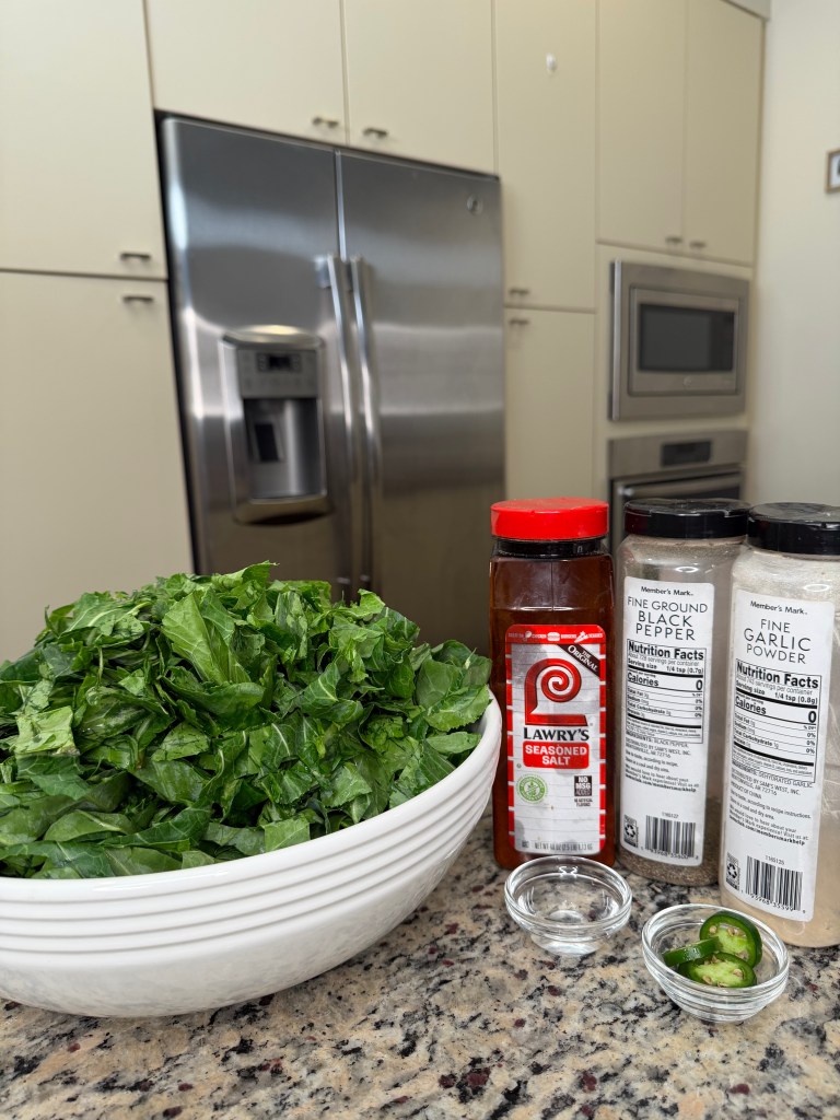 A bowl of chopped greens in a kitchen setting, with a refrigerator in the background. Seasoning containers, including Lawry's seasoned salt, black pepper, and garlic powder, are displayed alongside sliced jalapeños on a countertop.