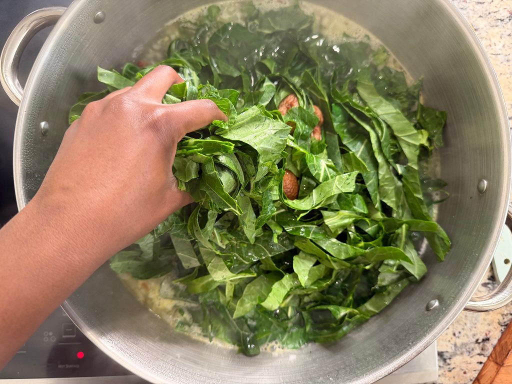 A hand adding fresh collard greens to a pot of boiling water, with almonds visible among the greens.