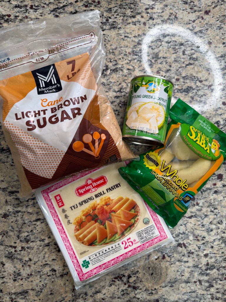A bag of light brown cane sugar, a can of young green jackfruit in brine, a package of spring roll pastry, and a bag of whole saba bananas placed on a granite countertop.