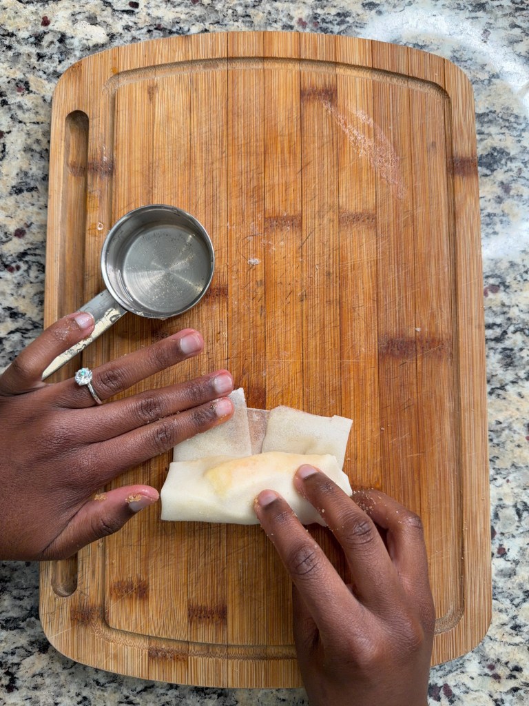 A person rolling a spring roll on a wooden cutting board, with a measuring cup nearby.