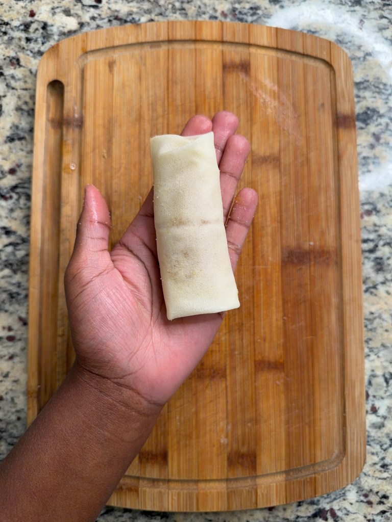 A person holding a spring roll on a wooden cutting board.