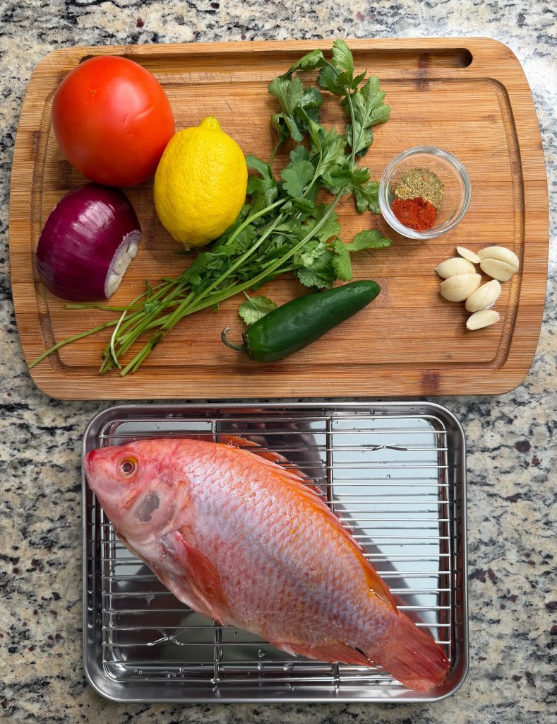 A large pink fish on a metal rack beside a wooden cutting board with a tomato, lemon, red onion, jalapeño, garlic cloves, fresh cilantro, and various spices.