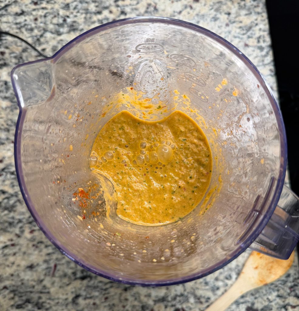 A blend of orange and green sauce inside a clear blender container, with bubbles and a textured appearance, placed on a granite countertop.