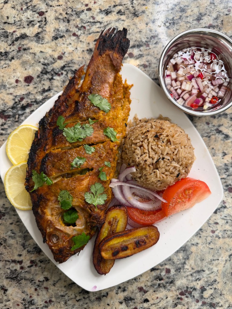 Grilled whole fish garnished with cilantro and lemon, served alongside seasoned rice, fried plantains, fresh tomato slices, and pickled onions in a bowl.