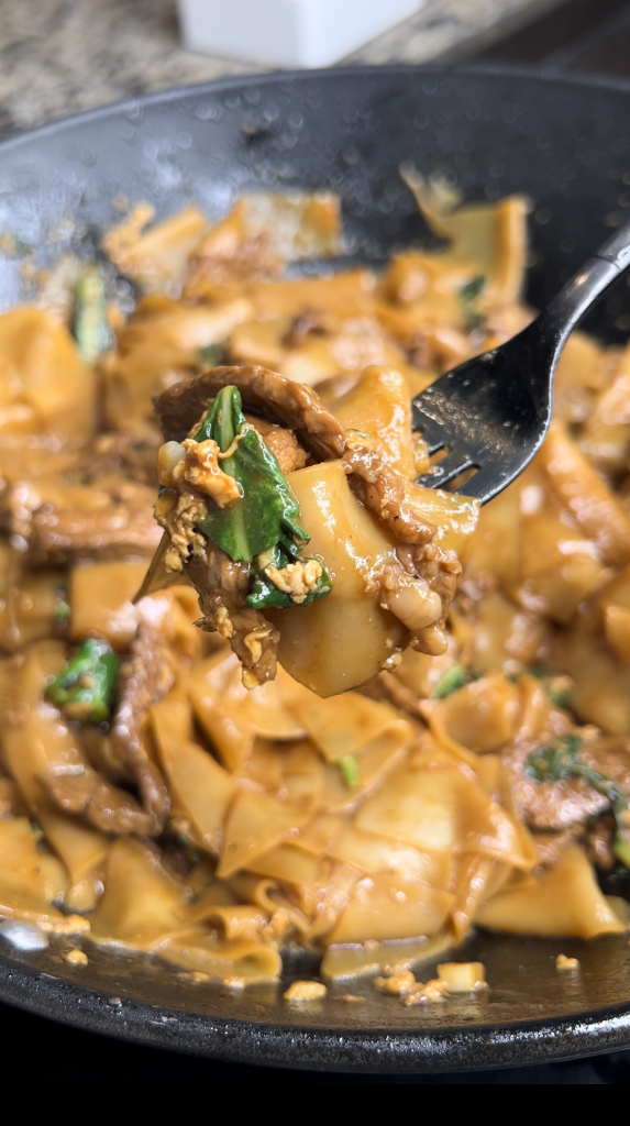 Close-up of a fork holding a portion of stir-fried beef and wide egg noodles with greens in a pan.