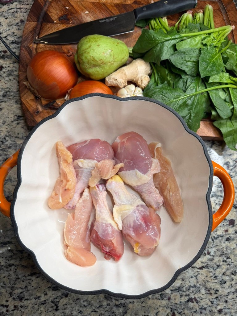 A bowl containing raw chicken pieces, surrounded by fresh vegetables including onions, a pear, ginger, garlic, and spinach on a wooden cutting board.