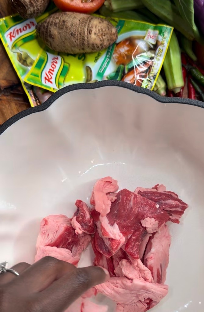 Raw meat pieces in a white bowl with various vegetables and seasonings in the background.