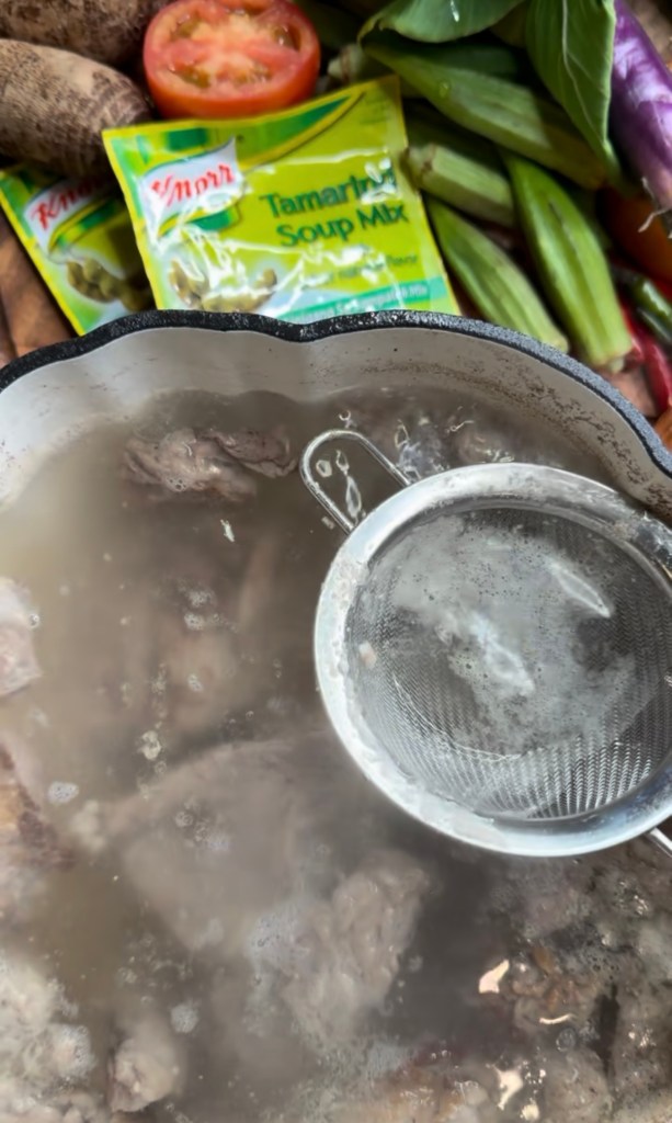 A pot of simmering broth with shredded meat and a strainer on the side, surrounded by fresh vegetables and a packet of tamarind soup mix.