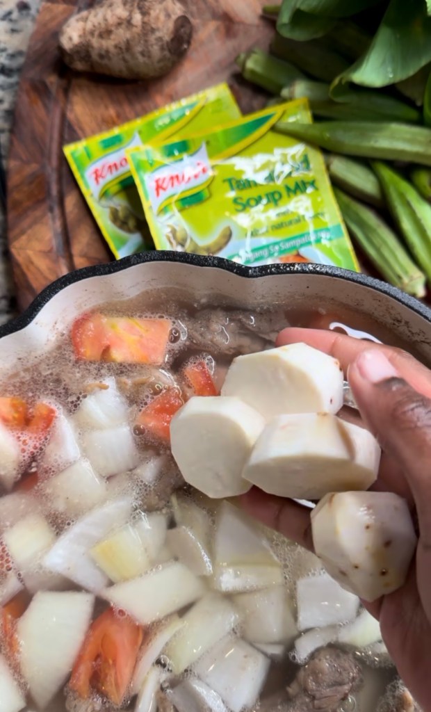 A hand holding sliced root vegetables above a pot of boiling soup that contains chopped onions, tomatoes, and meat, with packages of Knorr soup mix and okra in the background.