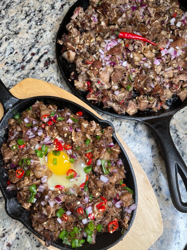 Two cast iron pans featuring savory dishes: one filled with chopped meat garnished with red onions and chilies, the other with meat and a cracked egg at the center surrounded by green onions and red chilies.