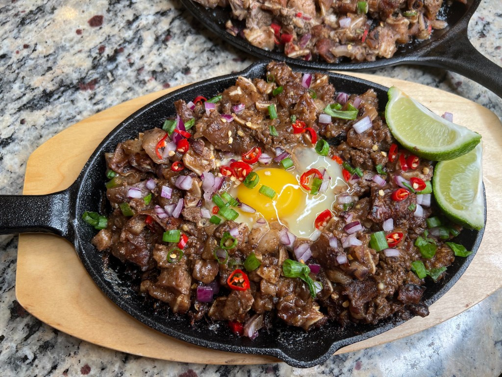 A hot sizzling plate of meat topped with red chili, green onions, and a raw egg in the center, accompanied by lime wedges.