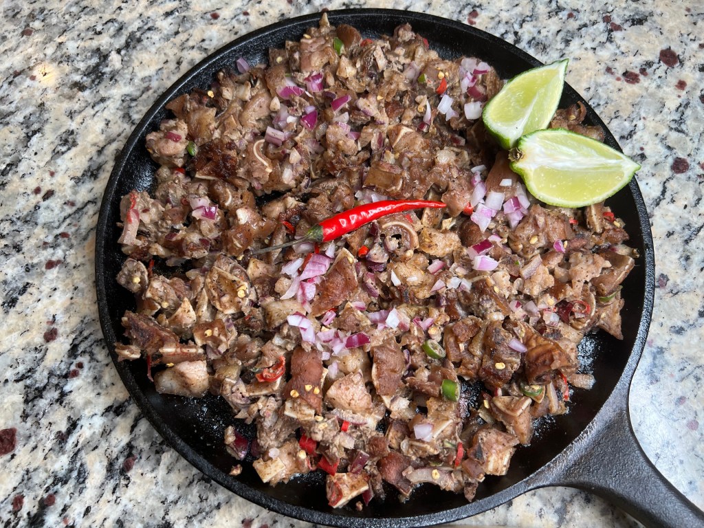 A cast-iron skillet filled with chopped, cooked meat, garnished with diced red onions, green chili peppers, and lime wedges on a granite countertop.