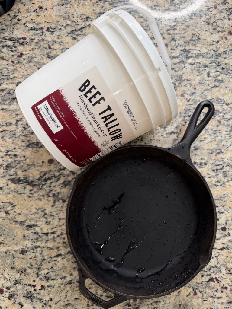 A bucket of beef tallow placed beside a cast iron skillet with a glossy layer of melted fat on its surface, set against a granite countertop.