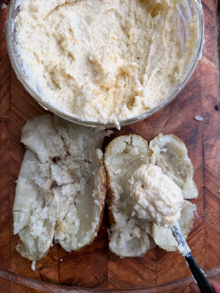 A wooden cutting board displaying two halved baked potatoes topped with a creamy mixture, alongside a bowl of the same mixture and a spoon.