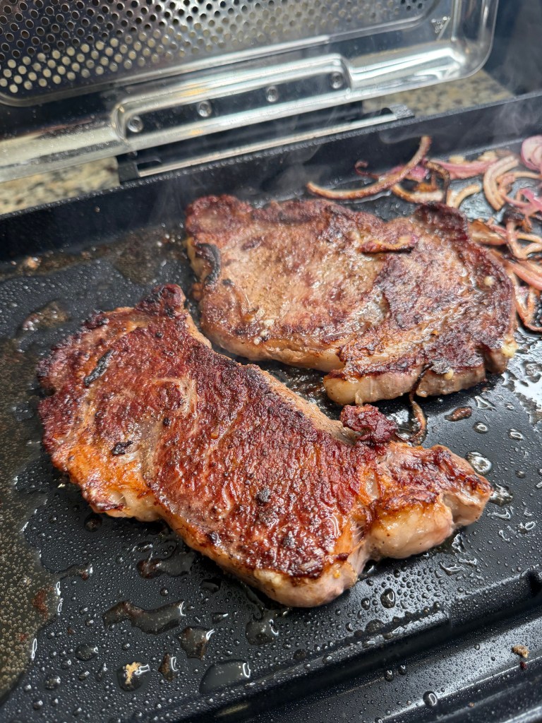 Two sizzling steaks cooking on a grill with onions in the background.