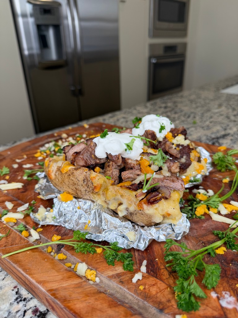 A loaded baked potato topped with steak, cheese, and dollops of sour cream, garnished with fresh parsley, on a wooden cutting board, with a modern kitchen in the background.