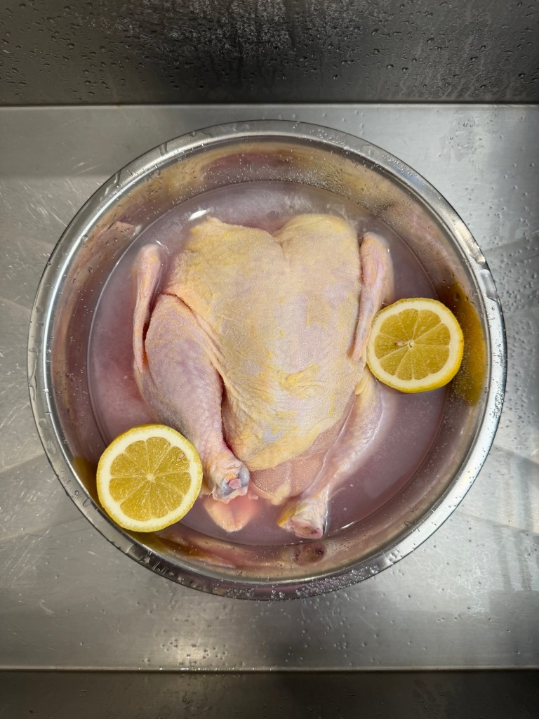 Raw chicken resting in a bowl of water with lemon slices, on a metal surface.