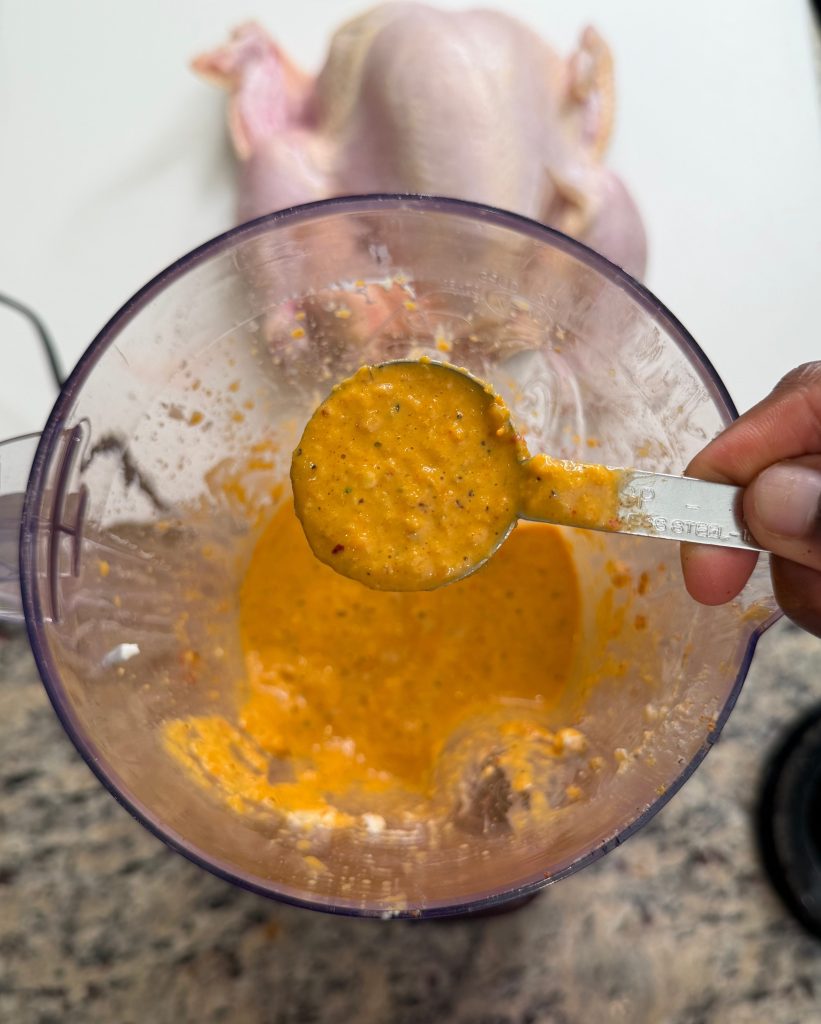 A measuring spoon filled with a thick, roasted honey garlic whole chicken marinade, held above a blender containing more of the same mixture, with a raw chicken in the background.