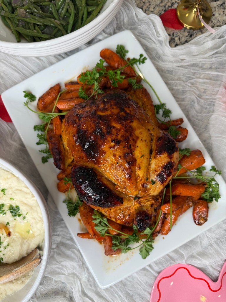 A beautifully roasted chicken garnished with fresh parsley, surrounded by caramelized carrots on a white platter. In the background, there is a bowl of green beans and a bowl of mashed potatoes.