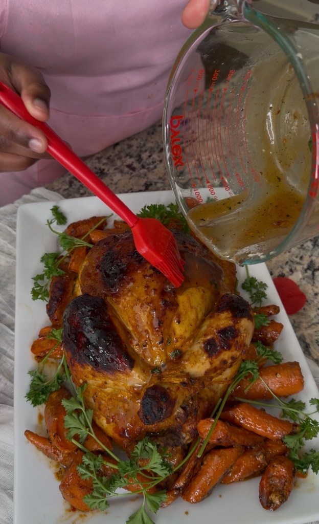 A person basting a roasted chicken with a brush, surrounded by glazed carrots and garnished with fresh parsley on a white platter.